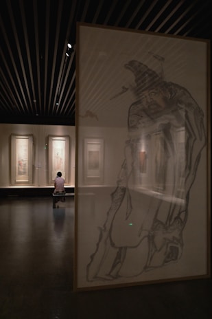 A dimly lit art gallery with large framed artworks on the walls. In the foreground, a detailed sketch of a person wearing traditional attire is partially visible. In the background, a visitor sits on a bench, observing the paintings.