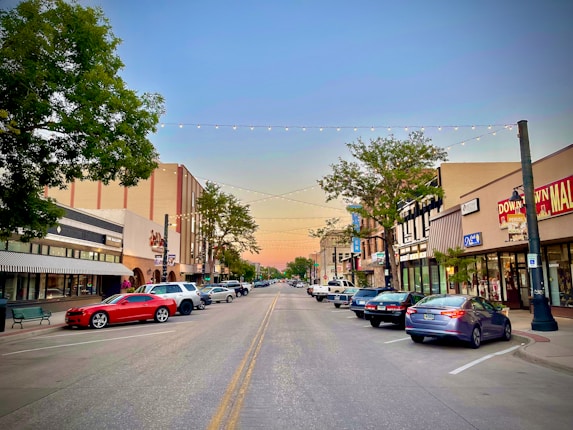 A small town street scene during sunset with cars parked along both sides. The street is adorned with hanging lights overhead and is flanked by buildings featuring shops and businesses. Trees line the sidewalks, and the sky shows a gradient from blue to orange.