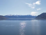 A calm lake reflecting snow-capped peaks and soft clouds on a clear day.