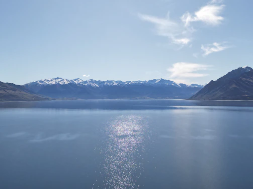 A calm ocean reflecting snow-capped mountains under a clear blue sky.