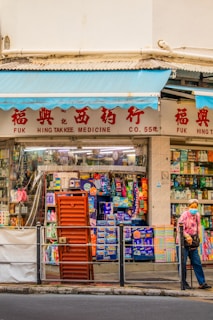 A street scene featuring a traditional medicine shop with red signage and a display of various products in the window. A person wearing a patterned shirt and a face mask stands outside, holding a walking stick. The storefront has blue awnings and a variety of colorful items visible through the glass.