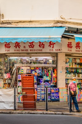 A street scene featuring a traditional medicine shop with red signage and a display of various products in the window. A person wearing a patterned shirt and a face mask stands outside, holding a walking stick. The storefront has blue awnings and a variety of colorful items visible through the glass.