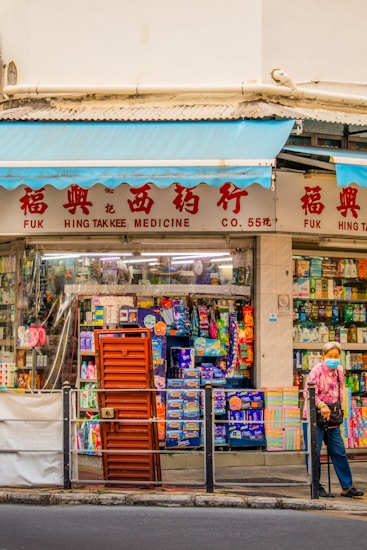 A street scene featuring a traditional medicine shop with red signage and a display of various products in the window. A person wearing a patterned shirt and a face mask stands outside, holding a walking stick. The storefront has blue awnings and a variety of colorful items visible through the glass.