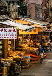 A bustling street market scene with various dried goods displayed in front of shops. Several people are browsing or managing the stalls, which are covered by striped awnings. A man in a mask is walking by, carrying a red bag. The environment is urban with visible signs featuring Asian characters, and the architecture is dense and gritty.