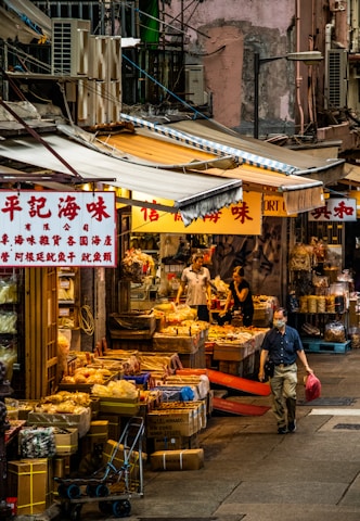 A bustling street market scene with various dried goods displayed in front of shops. Several people are browsing or managing the stalls, which are covered by striped awnings. A man in a mask is walking by, carrying a red bag. The environment is urban with visible signs featuring Asian characters, and the architecture is dense and gritty.