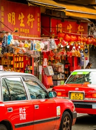 A bustling street scene with red taxis and a vibrant shop featuring numerous hanging decorations. The storefront displays bright signage with Chinese characters, while various trinkets, including lanterns and other decorative items, are visible.
