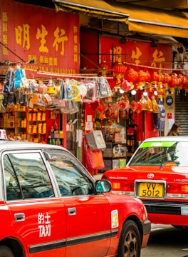A bustling street scene with red taxis and a vibrant shop featuring numerous hanging decorations. The storefront displays bright signage with Chinese characters, while various trinkets, including lanterns and other decorative items, are visible.