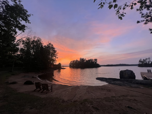 A tranquil lakeside scene at sunset with a vibrant sky featuring shades of pink, orange, and purple. Silhouetted trees line the shore while a solitary rock sits near a small dock. Two empty chairs are positioned on the sandy beach, facing the water, inviting relaxation and reflection.