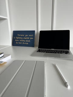 A workspace featuring a closed laptop, a white pen, a pad or tablet cover, stacked books, and a blue sign with an inspirational quote in yellow text. The background has white decorative wall paneling and the surface is a pristine white desk.