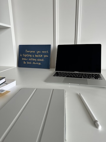 A workspace featuring a closed laptop, a white pen, a pad or tablet cover, stacked books, and a blue sign with an inspirational quote in yellow text. The background has white decorative wall paneling and the surface is a pristine white desk.