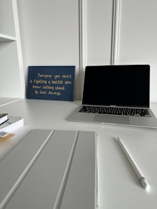 A workspace featuring a closed laptop, a white pen, a pad or tablet cover, stacked books, and a blue sign with an inspirational quote in yellow text. The background has white decorative wall paneling and the surface is a pristine white desk.