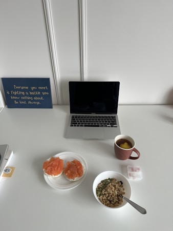 A clean white desk features a closed laptop, a motivational sign on the left, and a breakfast setting. The breakfast includes two slices of bread with salmon on a plate, a bowl of granola with seeds and nuts, a cup of tea with lemon in a red mug, and a pill organizer. The sign reads 'Everyone you meet is fighting a battle you know nothing about. Be kind. Always.'