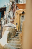 A rustic, futuristic dwelling built into a rocky landscape with weathered stone steps leading up to a round, metallic door. Vegetation grows sporadically on the rocks, and various adornments hang near the entrance, suggesting a blend of technology and nature.