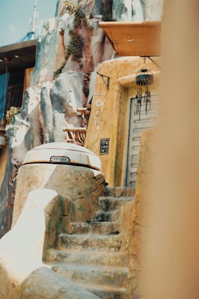 A rustic, futuristic dwelling built into a rocky landscape with weathered stone steps leading up to a round, metallic door. Vegetation grows sporadically on the rocks, and various adornments hang near the entrance, suggesting a blend of technology and nature.