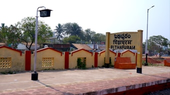 A railway station platform with a sign displaying the name Pithapuram in three languages. The area is surrounded by trees, with a low wall decorated in yellow and red. There are streetlights and a few scattered houses with slanted roofs visible in the background.