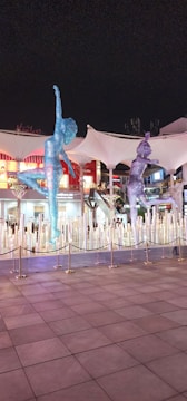 Two large, glittering sculptures of athletes are prominently displayed in an outdoor area. The figures, composed of a myriad of sparkling lights, appear dynamic and are positioned as if mid-motion. Behind the sculptures, a series of illuminated buildings and a canopy can be seen, along with a cordoned off area.