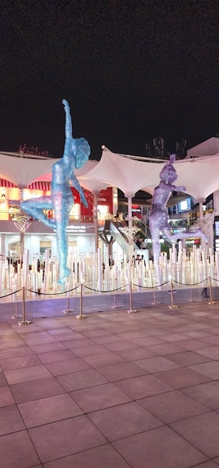 Two large, glittering sculptures of athletes are prominently displayed in an outdoor area. The figures, composed of a myriad of sparkling lights, appear dynamic and are positioned as if mid-motion. Behind the sculptures, a series of illuminated buildings and a canopy can be seen, along with a cordoned off area.