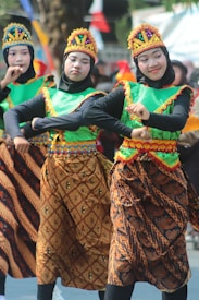Three individuals are dressed in traditional costumes with vibrant colors and intricate patterns. They wear headgear decorated with colorful ornaments and are performing a dance. Their attire features green tops with orange and blue trim, and brown batik-style skirts with elaborate designs.