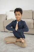 A child sits cross-legged on a carpeted floor with eyes closed and hands in a prayer position. The background includes a beige sofa with a white pillow. The child is wearing a dark long-sleeve shirt and beige pants, appearing peaceful and focused.