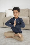 A child sits cross-legged on a carpeted floor with eyes closed and hands in a prayer position. The background includes a beige sofa with a white pillow. The child is wearing a dark long-sleeve shirt and beige pants, appearing peaceful and focused.