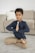 A child sits cross-legged on a carpeted floor with eyes closed and hands in a prayer position. The background includes a beige sofa with a white pillow. The child is wearing a dark long-sleeve shirt and beige pants, appearing peaceful and focused.