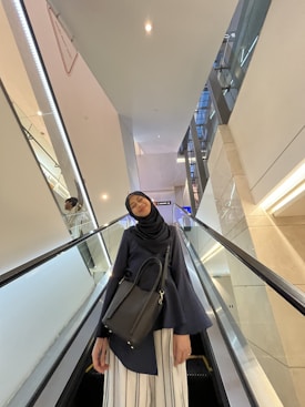 A person wearing a hijab and a dark blue outfit stands on an escalator in what appears to be a modern indoor space. They are holding a black bag and are positioned slightly off-center. The background includes the escalator's glass sides, white walls, and some reflective panels.