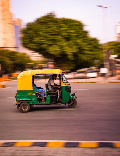 A blurred image of a green and yellow auto rickshaw driving on a city street. The background includes large trees and some urban buildings, creating a motion effect as the vehicle speeds by.