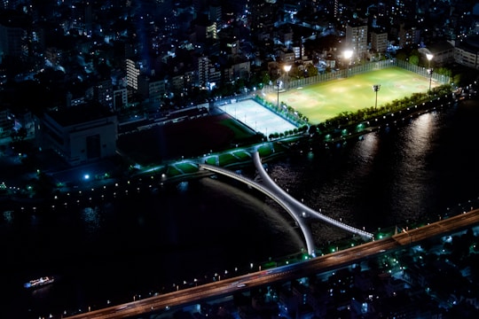 An aerial night view of a cityscape featuring a well-lit sports complex with green fields and a blue-lit sports court, surrounded by urban buildings. A distinctive bridge crosses a river that reflects the lights from the surrounding structures.