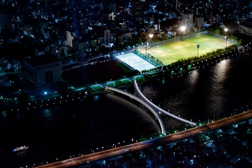 An aerial night view of a cityscape featuring a well-lit sports complex with green fields and a blue-lit sports court, surrounded by urban buildings. A distinctive bridge crosses a river that reflects the lights from the surrounding structures.