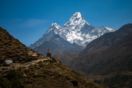 A magnificent snow-capped mountain rises majestically against a clear blue sky, with rugged, forested terrain in the foreground. A small stupa sits along a path that navigates the hills, adding a spiritual element to the natural landscape.