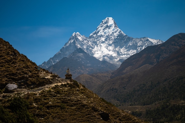A magnificent snow-capped mountain rises majestically against a clear blue sky, with rugged, forested terrain in the foreground. A small stupa sits along a path that navigates the hills, adding a spiritual element to the natural landscape.
