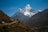 A magnificent snow-capped mountain rises majestically against a clear blue sky, with rugged, forested terrain in the foreground. A small stupa sits along a path that navigates the hills, adding a spiritual element to the natural landscape.
