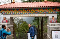 A man with a blue backpack smiles while standing under a colorful gate labeled Pasang Lhamu Gate. Another person in a blue jacket is on the left, raising a hand in gesture. The gate is adorned with statues of women on both sides and features vibrant colors such as red, blue, and yellow. The background reveals a lush forest setting.