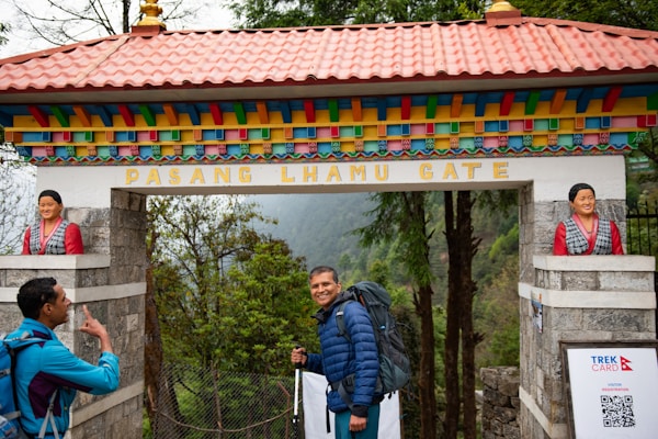 A man with a blue backpack smiles while standing under a colorful gate labeled Pasang Lhamu Gate. Another person in a blue jacket is on the left, raising a hand in gesture. The gate is adorned with statues of women on both sides and features vibrant colors such as red, blue, and yellow. The background reveals a lush forest setting.