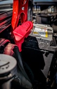 A close-up view of a car battery with red jumper cables attached. The scene captures intricate details of the battery terminals and cables, with a focus on the metal clamps and connectors. The surrounding area of the engine compartment is partially visible, carrying a utilitarian and mechanical feel.