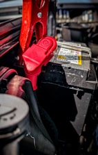 A close-up view of a car battery with red jumper cables attached. The scene captures intricate details of the battery terminals and cables, with a focus on the metal clamps and connectors. The surrounding area of the engine compartment is partially visible, carrying a utilitarian and mechanical feel.