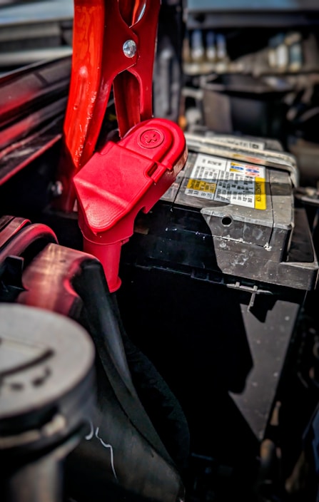 A close-up view of a car battery with red jumper cables attached. The scene captures intricate details of the battery terminals and cables, with a focus on the metal clamps and connectors. The surrounding area of the engine compartment is partially visible, carrying a utilitarian and mechanical feel.