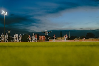 A football game taking place under a darkening evening sky with stadium lights illuminating the field. Players are scattered across the field, wearing different colored uniforms. A referee stands with arms outstretched, possibly signaling a decision. The scoreboard and goalposts are visible in the background.