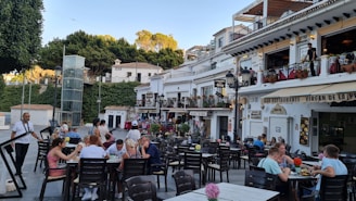 A bustling outdoor restaurant scene with multiple tables occupied by people enjoying meals and conversations. The setting features a charming European-style facade with white buildings, balconies adorned with plants, and various signage. Trees and greenery encircle the area, adding to the lively atmosphere.