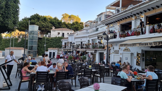 A bustling outdoor restaurant scene with multiple tables occupied by people enjoying meals and conversations. The setting features a charming European-style facade with white buildings, balconies adorned with plants, and various signage. Trees and greenery encircle the area, adding to the lively atmosphere.