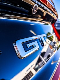 A close-up of a car's rear section featuring a shiny metallic GT badge. The background includes blurred reflections and vibrant lighting effects, hinting at an urban setting.