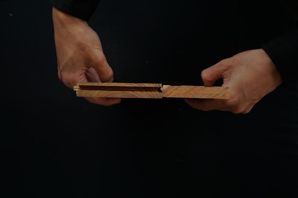 Two hands are holding wooden planks with a tongue and groove design against a dark background.