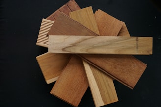 A collection of wooden samples arranged in a circular pattern on a black background. The samples are various shades of brown, with some displaying distinctive wood grain patterns and textures.