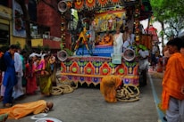 A colorful religious parade featuring a decorated chariot adorned with flowers and intricate patterns. Several people dressed in traditional attire are present, including a person lying prostrate on the ground in front of the chariot. The surrounding crowd is engaged in the processional activities.