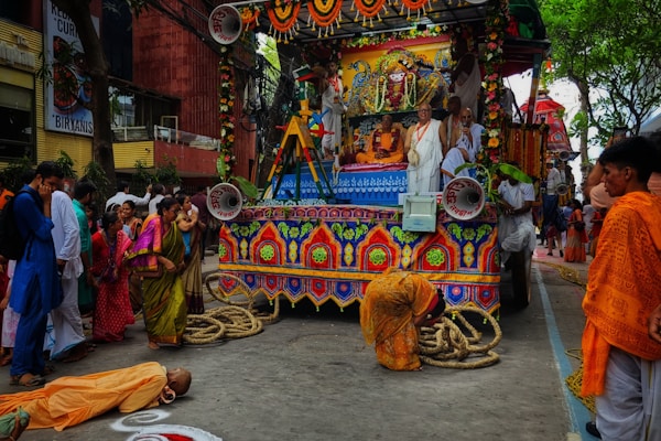 A colorful religious parade featuring a decorated chariot adorned with flowers and intricate patterns. Several people dressed in traditional attire are present, including a person lying prostrate on the ground in front of the chariot. The surrounding crowd is engaged in the processional activities.