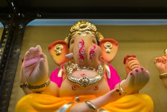 A colorful statue of a deity with an elephant head, adorned with intricate jewelry and bright clothing. The figure has multiple hands, one of which is raised in a blessing gesture. The details include a headdress, ornate designs on the body, and vivid accents in pink and orange.
