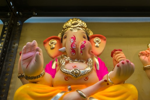 A colorful statue of a deity with an elephant head, adorned with intricate jewelry and bright clothing. The figure has multiple hands, one of which is raised in a blessing gesture. The details include a headdress, ornate designs on the body, and vivid accents in pink and orange.