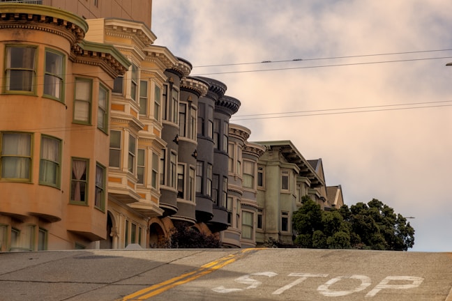 A row of classic San Francisco-style bay window buildings line a steep street, featuring various beige, cream, and gray tones. The foreground displays the word 'STOP' painted on the ascending road. Overhead, power lines stretch across the partly cloudy sky.