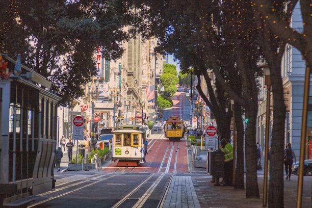 A bustling city street features iconic cable cars traveling down tracks lined with trees and historic buildings on a sunny day. American flags hang from some buildings amid various shop signs and bustling pedestrian activity.