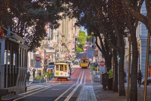A bustling city street features iconic cable cars traveling down tracks lined with trees and historic buildings on a sunny day. American flags hang from some buildings amid various shop signs and bustling pedestrian activity.